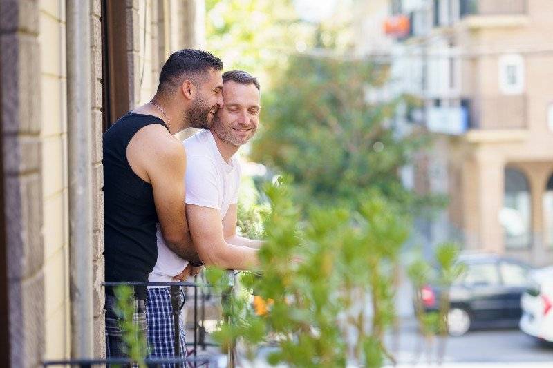 A gay couple embraces and smiles on a leafy city balcony, enjoying a peaceful moment together in a sunny, urban LGBTQ+ destination.