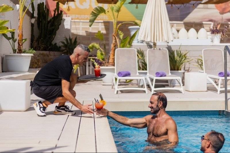 A shirtless man in the pool receives a colorful cocktail from staff at The Purple Hotel Ibiza, a gay-only adults resort with fun vibes.

