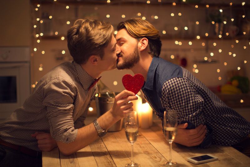 Dois homens se beijando sobre uma mesa de madeira, segurando um coração vermelho, com duas taças de champanhe sobre a mesa e luzes de fadas atrás