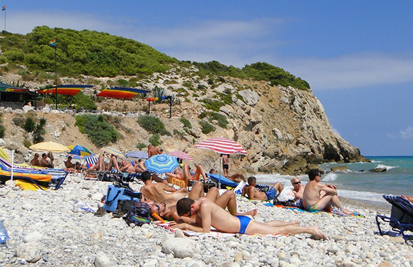 Crowded gay nude beach with rainbow flags, sunbathers, and umbrellas on a pebbly shoreline.