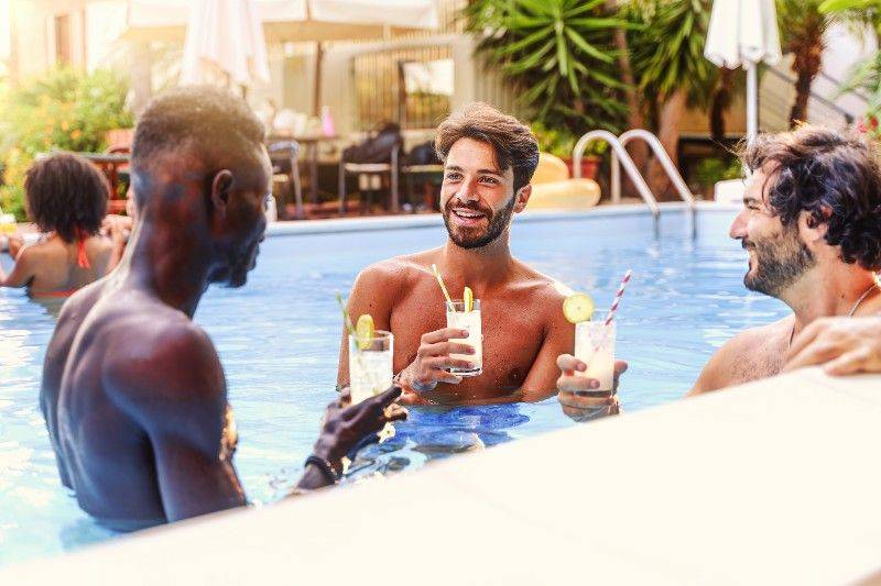 Vibrant gay resort pool party in Las Vegas, with rainbow flags, palm trees, and smiling men enjoying drinks and sunshine.