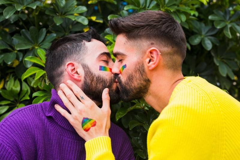 Two men share a tender kiss while wearing bright sweaters (one purple, one yellow), with rainbow flags painted on their cheeks and hands, set against a leafy green background.