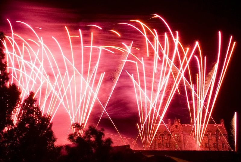 Fogos de artifício vermelhos e rosa brilhantes iluminam o céu noturno acima de um edifício histórico com árvores em silhueta no primeiro plano.