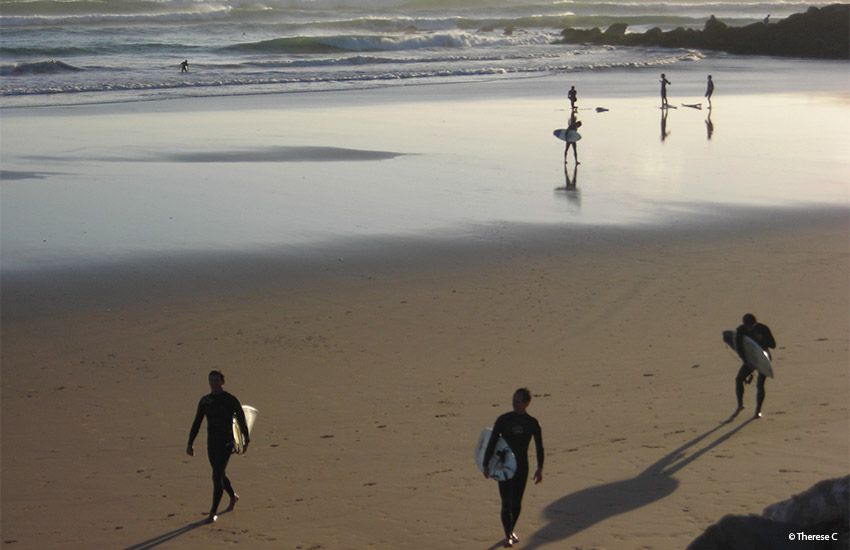 Surfers walking along the wet sand at a wide, calm gay beach perfect for naturists and sports lovers.