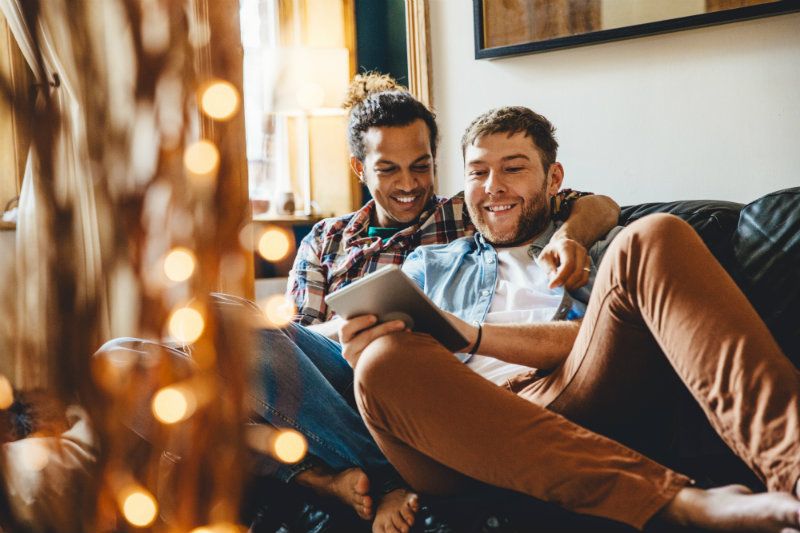 A happy couple sits on a couch, one holding a tablet as they laugh together in a cozy room, softly illuminated by string lights in the foreground.