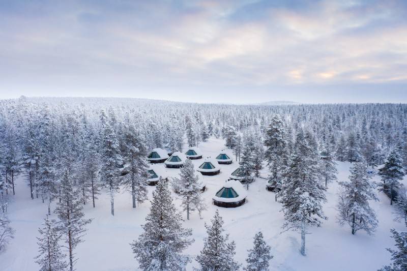 A winter resort with glass-roofed cabins surrounded by snow-covered trees under a pastel sky.