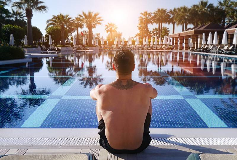 Homem com tatuagem nas costas sentado à beira da piscina, de frente para as palmeiras ao pôr do sol.