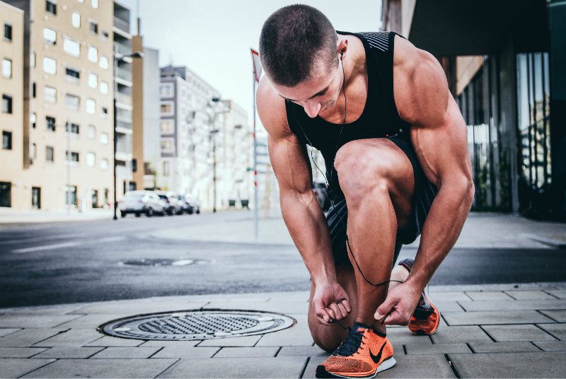 A muscular man in athletic gear ties his running shoes on an urban sidewalk, preparing for a workout with focused determination.