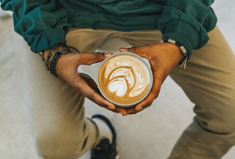 A close-up of hands holding a latte in a ceramic cup, featuring intricate heart-shaped latte art, against a backdrop of casual attire and warm tones.