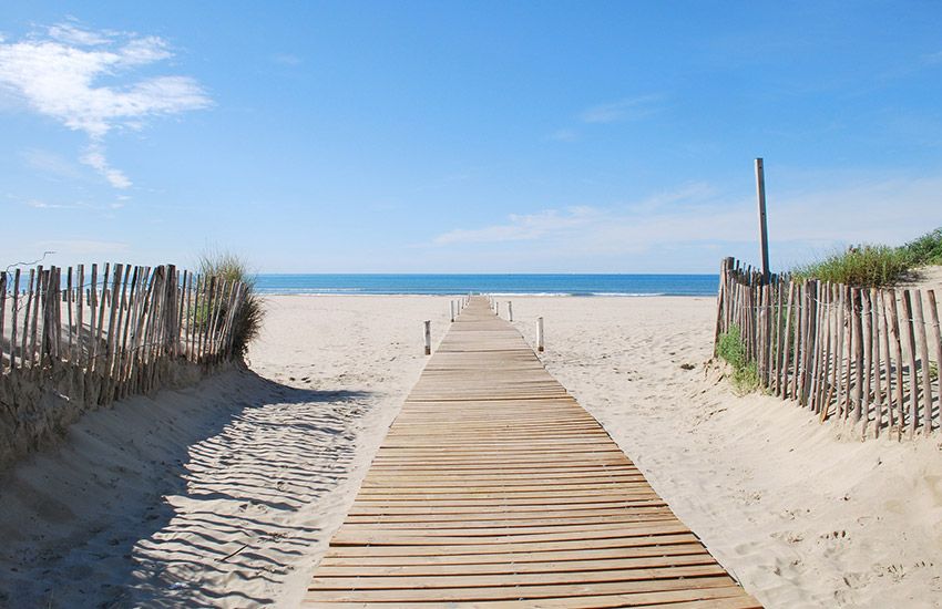 Wooden boardwalk leading to a wide, pristine sandy gay beach with blue sky and calm Mediterranean waters.