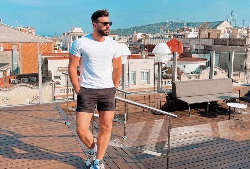 A man in sunglasses and shorts standing on a rooftop terrace with a cityscape in the background.