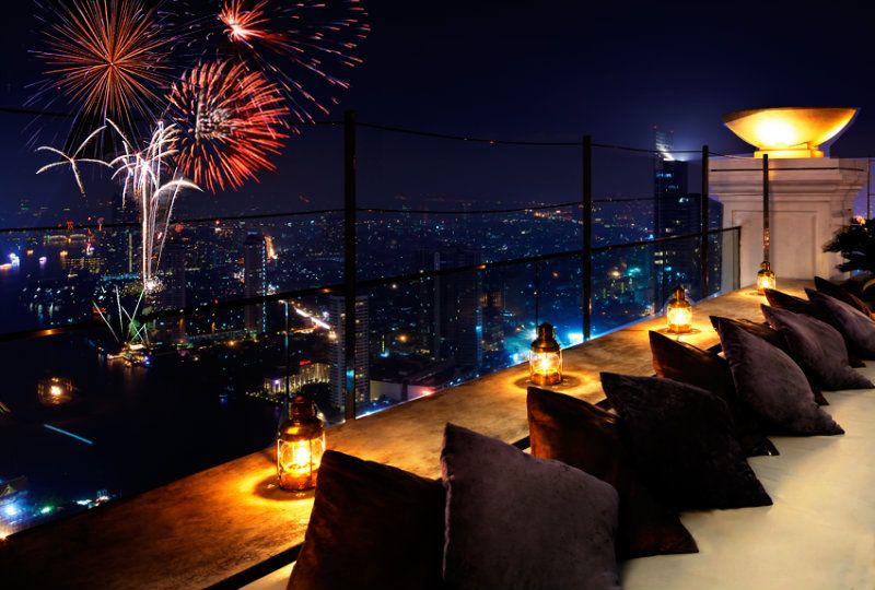 Vista de um terraço na cobertura com assentos aconchegantes e lanternas, com vista para o horizonte de uma cidade iluminada por fogos de artifício vibrantes em um céu noturno escuro.