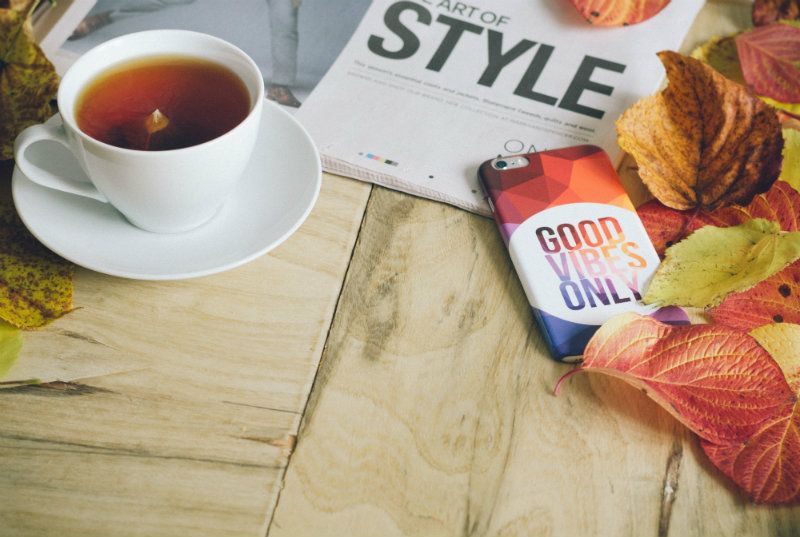 A cup of tea sits on a wooden table alongside autumn leaves, a smartphone with a