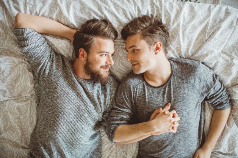 Two men lying on a bed, facing each other and holding hands, sharing an affectionate moment with soft smiles and cozy, matching gray sweaters.