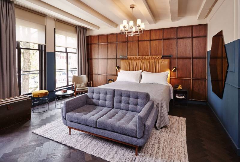 A stylish hotel room with a modern gray sofa, a cozy bed, wooden panel walls, and large windows with natural light.