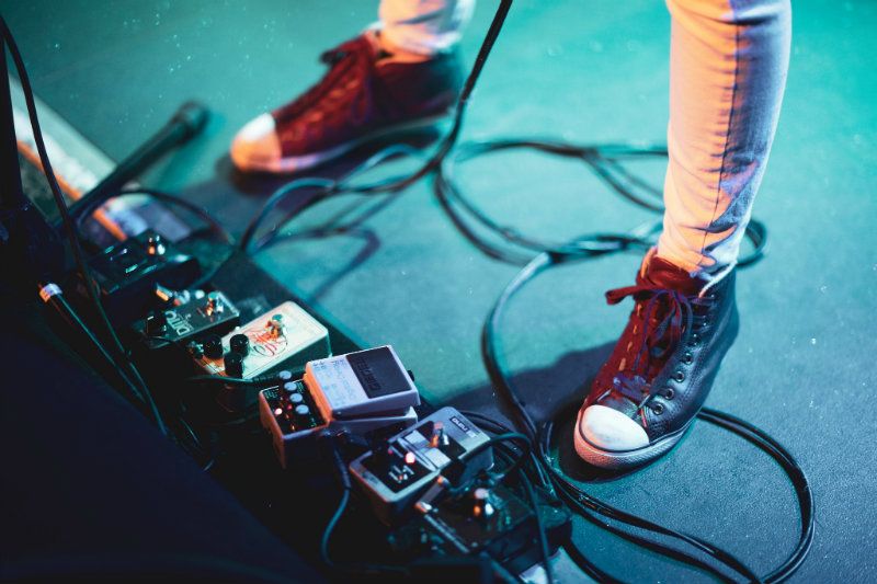 Close-up dos pés de um músico usando tênis preto de cano alto em um palco cercado por cabos e pedais de efeitos de guitarra, com iluminação colorida no palco.